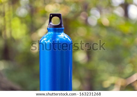 Metal water bottle in green forest over blurred background, hiking and sport equipment