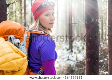 Woman Hiking In White Winter Forest, Backlight By Morning Sunlight Rays, Recreation And Healthy Lifestyle Outdoors In Nature. Beauty Blond Hiker Backpacker Looking At Camera On Sunset.