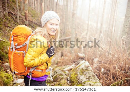 Woman hiking in autumn forest in mountains. Trekking, recreation and healthy lifestyle outdoors in nature. Beauty blond backpacker looking at camera smiling, bright light sunlight in background.