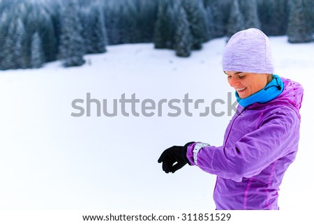Woman runner checking sports watch on winter run. Sport, fitness inspiration and motivation. Young happy woman cross country running in mountains on snow winter. Female trail runner jogging outdoors.