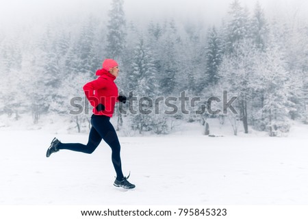 Woman running on snow in winter mountains. Sport, fitness inspiration and motivation. Young happy woman trail running in mountains on snow, winter day. Female trail runner jogging outdoors.