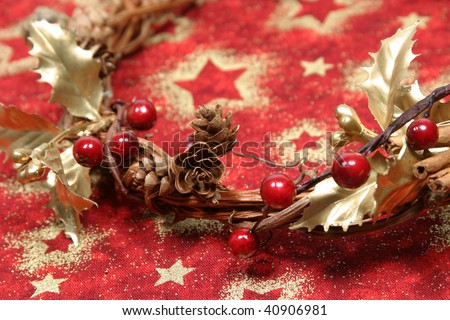 Christmas wreath with golden holly leaves, cones, red berries and cinnamon on red textile with gold stars. Shallow DOF