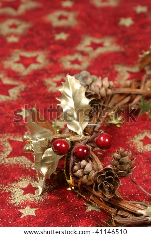Christmas wreath with golden holly leaves, cones, red berries and cinnamon on red textile with gold stars. Copy space, shallow DOF