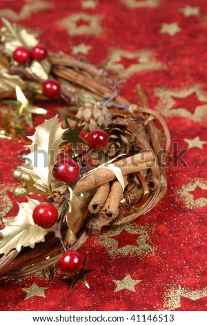 Christmas wreath with golden holly leaves, cones, red berries and cinnamon on red textile with gold stars. Shallow DOF