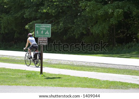 Woman cyclist riding in a public park - signs posted for the all-purpose trail.  Warning to keep pets leashed.  Room at right for text.