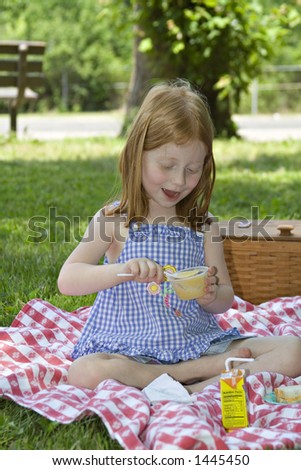 Small red-haired girl eating a cup of applesauce outdoors in a park - picnic setting.