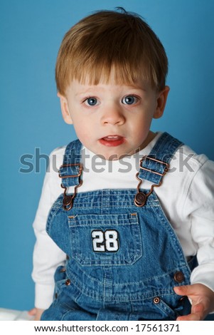 Boy toddler age 2 in bib overalls, blue background.