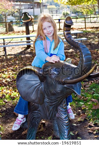 A small redhead girl sitting on an elephant statue in a public park.