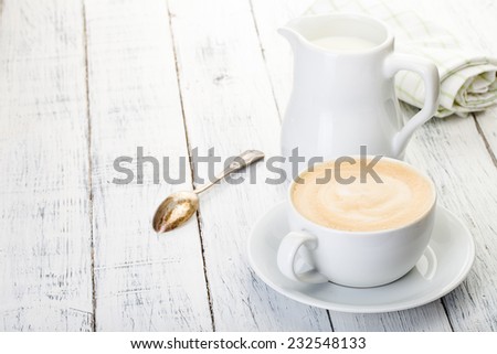 cup of cappuccino and jug of milk on old painted white wooden table.