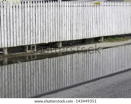 White picket fence reflected by puddle along pavement on city street