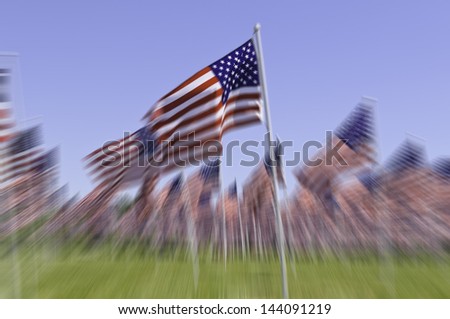 Radial blur of many American flags standing together, with foreground flag prominent, on green lawn under blue sky