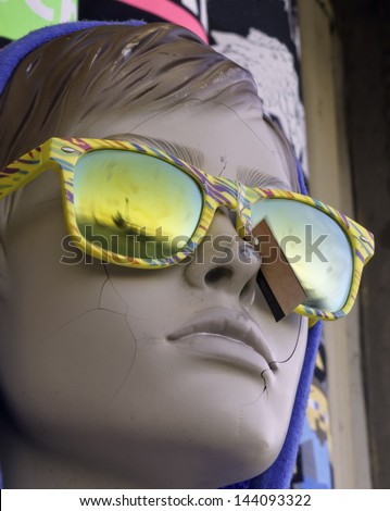 Portrait of serene mannequin with cracked face, eyes hidden behind tropical sunglasses with colored frames and blurred lenses, blank tag by her nose, staring at ocean beach