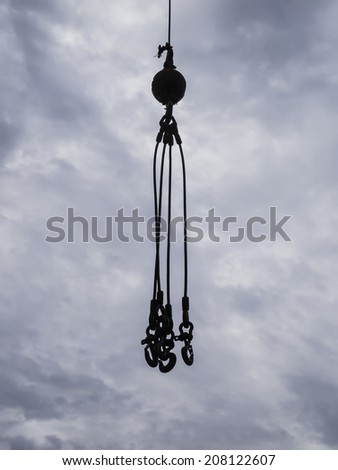 Industrial equipment in silhouette: Four hooks to lift commercial fishing tackle and other heavy objects on ship or shore hang in mid air against a cloudy sky