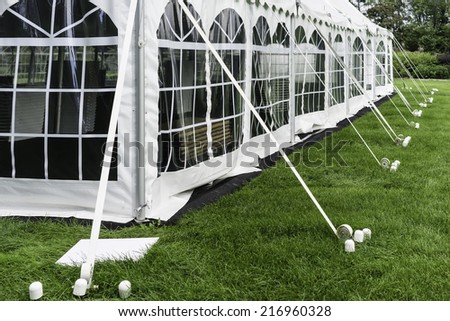 Corner and side of large white event tent with plastic windows, anchored on garden lawn in summer