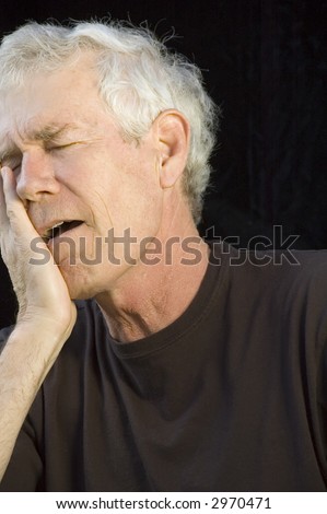 Mature white man with gray hair, eyes closed, head in right hand, mouth open, with expression of pain or regret or dismay