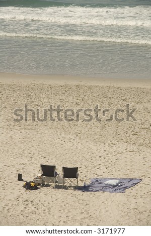 Pair of folding chairs, blanket, and other personal belongings left unattended on beach with many footprints near ocean waves