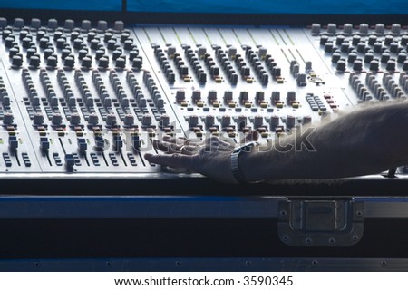 Technician\'s left hand makes adjustment on audio control panel in booth at outdoor concert