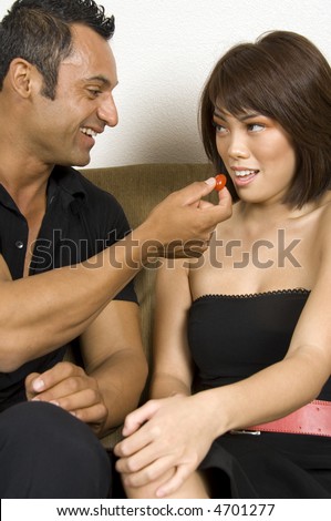 Grinning Hispanic man offers cherry tomato to pretty young Asian-American woman with bare shoulders