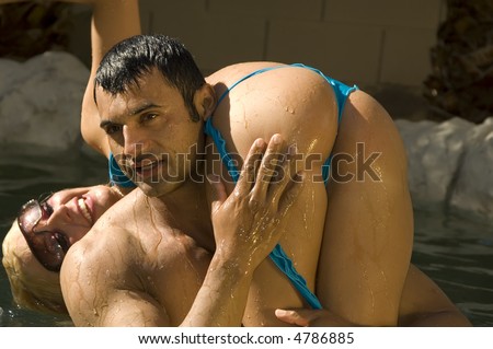 Strong wet Hispanic man holds grinning blond woman in bikini upside down over one shoulder, hand on her leg, in backyard swimming pool