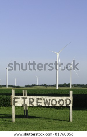 Irony of rural power sources: in foreground, road sign indicating firewood for sale; in background, group of wind turbines (turbine blades stand still in absence of wind) (copy space in sky)
