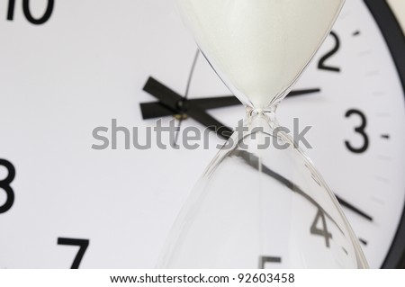 Two measurements of time: White sand falling inside hourglass, with round analog clock in  background (focus on neck of hourglass), shallow depth of field