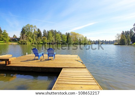 Lake spring waterfront with pier and two blue chairs.