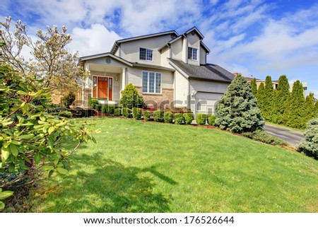 Two story siding house with brick trim, column porch and garage. Green lawn with trimmed hedges, fir trees and flourishing trees