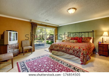 Contrast color bedroom with walkout deck. Room furnished with antique iron frame bed, nightstands, cabinet with tv and old fashioned chairs.