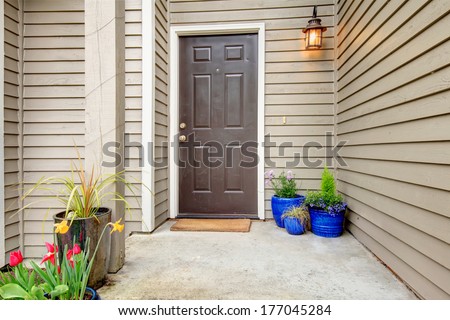 Open Porch With Concrete Floor, Column And Entrance Brown Door. Decorated With Hanging Lantern Light And Pots With Flowers
