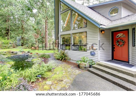 Beautiful house with orange entrance door, glass wall and picturesque man made pond with flower pots and wooden chairs