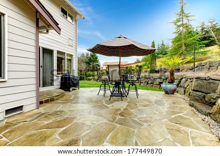 Concrete floor cozy patio area with iron table set and patio umbrella. Patio area surrounded by green terrace landscaping