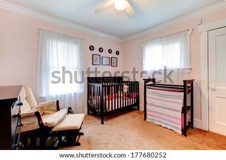 Bright nursery room with light pink walls, carpet floor. Furnished with dark brown wooden crib, chair and dresser