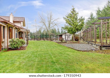 Farm house backyard with a lawn, trees and wooden grids. View of the fenced horse barn