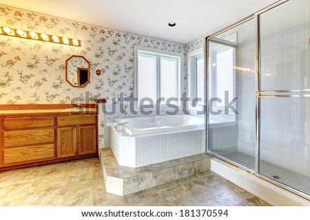 Spacious bathroom with floral wallpaper, concrete floor and windows. View of white  tub, glass door shower and wooden cabinets