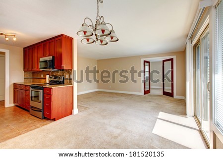 Empty bright living room with carpet floor. View of small kitchen area with wooden cabinets and steel appliances