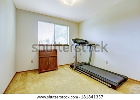 Bright gym room with one window and carpet floor. Running exercise equipment and wooden cabinet