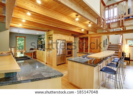 View of shiny kitchen with steel appliances, light wooden cabinets and ceiling beams. View of bar with stools and counter top with sink