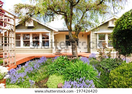 Clapboard siding house exterior. View of wooden deck and flower bed with blooming plants and trees