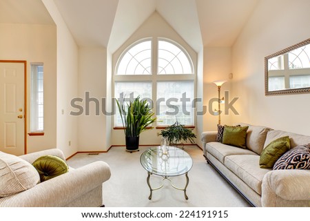 Luxury family room in soft creamy tones with high ceiling and arch window. Room with sofa and armchair, glass top coffee table. Room decorated with green plants