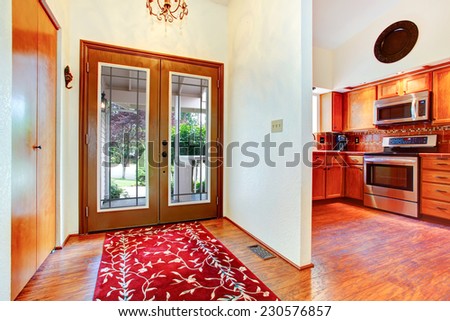 House interior. Entrance hallway with glass door, hardwood floor and bright red rug. View of kitchen room with orange cabinets and steel stove