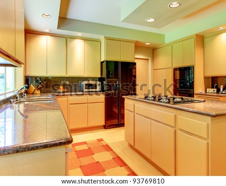Large kitchen with honey wood and black appliances and red rug.