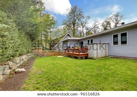 Back yard grass and bushes of small grey house wth simple deck. spring day