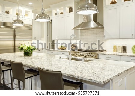 White kitchen design features large bar style kitchen island with granite countertop illuminated by modern pendant lights. Stainless steel appliances framed by white shaker cabinets . Northwest, USA