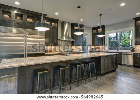 Modern kitchen with brown kitchen cabinets, oversized kitchen island with bar stools, granite countertops, huge refrigerator and beige backsplash. Northwest, USA