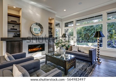 Chic living room filled with built-in cabinets flanking round mirror atop grey tile fireplace, tufted sofa facing two armchairs and window wall overlooking  lush outdoors. Northwest, USA