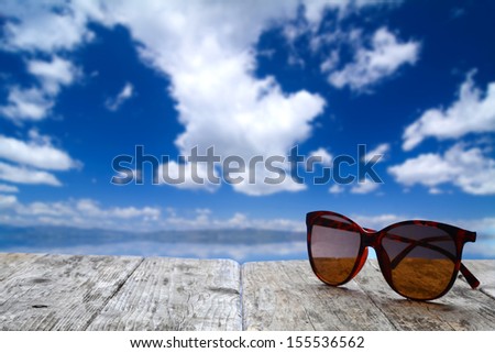 Sunglasses on a wood platform (boat dock or board walk) on a bright sunny day with blue sky and water background.