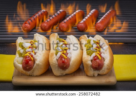 Three Hot Dogs with Mustard, Ketchup, pickle relish and onions and Hot Dogs on a Hot Flaming Barbecue Grill in the Background.