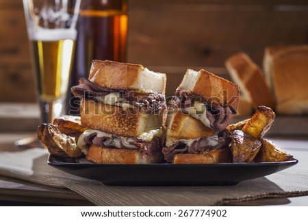 Sliced roast beef and melted cheese sandwich on a black plate with wedge cut rosemary potato fries and beer