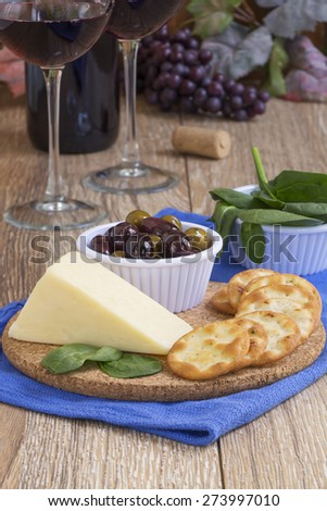 A rustic wooden table setting with a wedge of white cheddar cheese,crackers,kalamata olives, spinach leaves with two glasses of red wine, a wine bottle and wine bottle cork