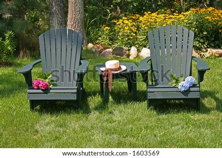 two wooden empty lawn chairs are in a garden. Woman\'s hat is on the table. Red and blue flowers are on the chairs